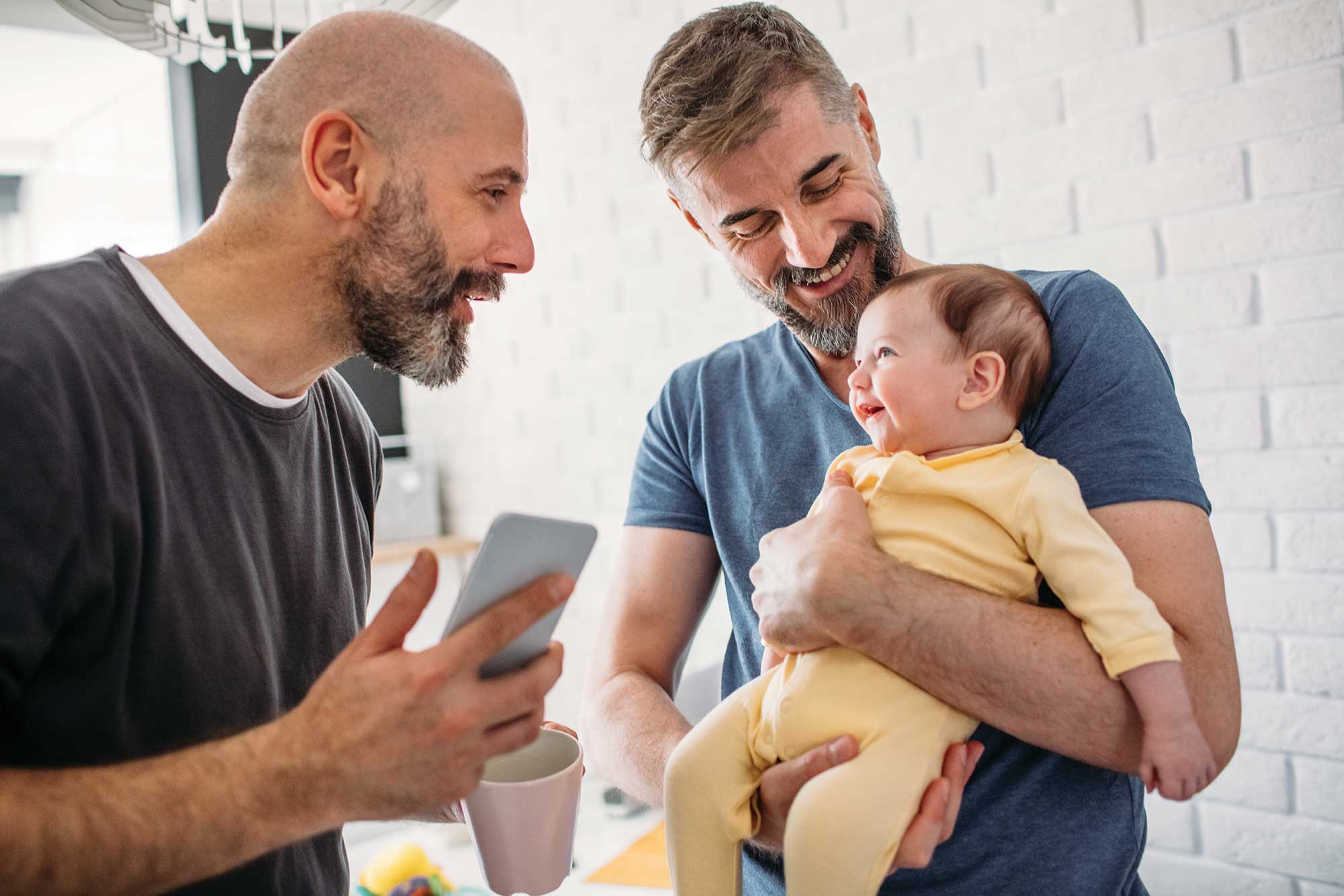 One man holding a cell phone, one man holding a baby, happy family